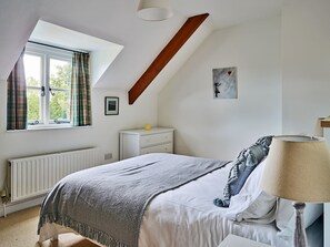 Ein Schlafzimmer mit schrägem Dach, einem Bett mit grauer Decke, einer Nachttischlampe, einer Kommode und einem Fenster mit Vorhängen.