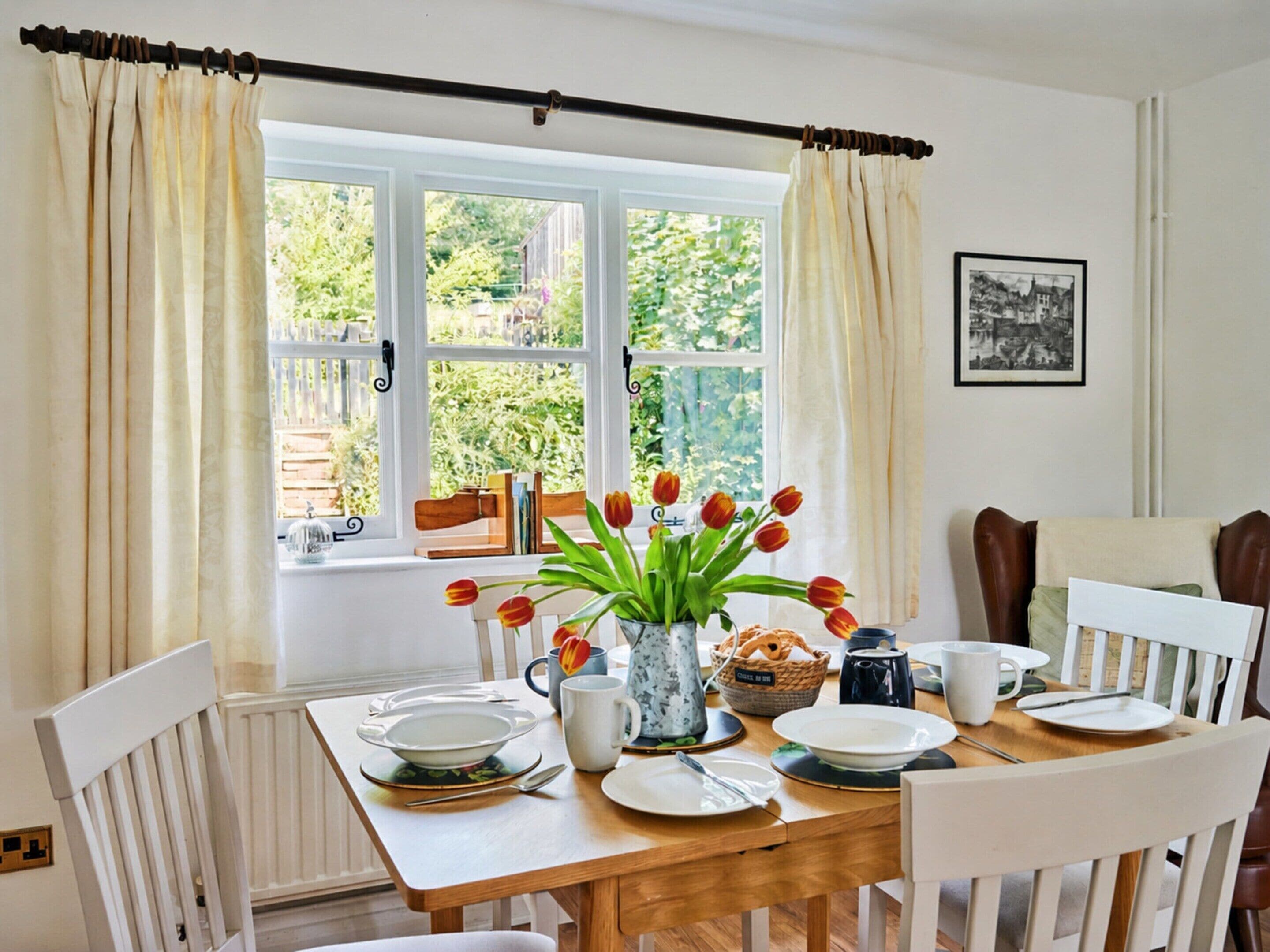 Ein Essbereich mit Tisch für vier Personen, einem fensterbelaubten Fenster, einer Vase mit Tulpen und einem gerahmten Bild an der Wand.