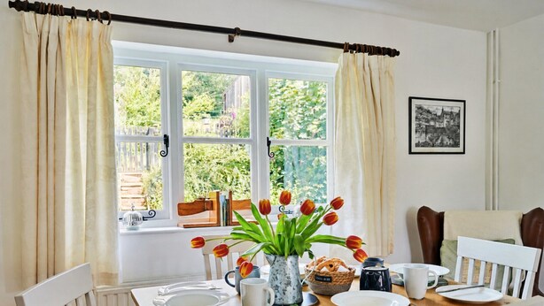 A dining area with a table set for four, a window with curtains, a vase with tulips, and a framed picture on the wall. - Historic Cornish Valley Cottage for 5 (Crediton)
