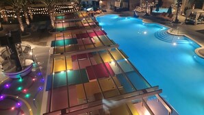 Une piscine avec un espace de détente, des palmiers et des guirlandes lumineuses.