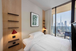 Ein Hotelzimmer mit einem Bett, einer Wandablage, einer roten Lampe, einem gerahmten Kunstwerk und Blick auf die Stadt durch große Fenster.