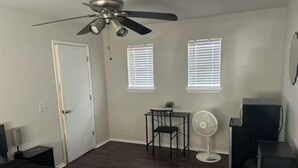 Een slaapkamer met een bed, een plafondventilator, een bureau, een stoel, een klein tafeltje, een ventilator en een dressoir.