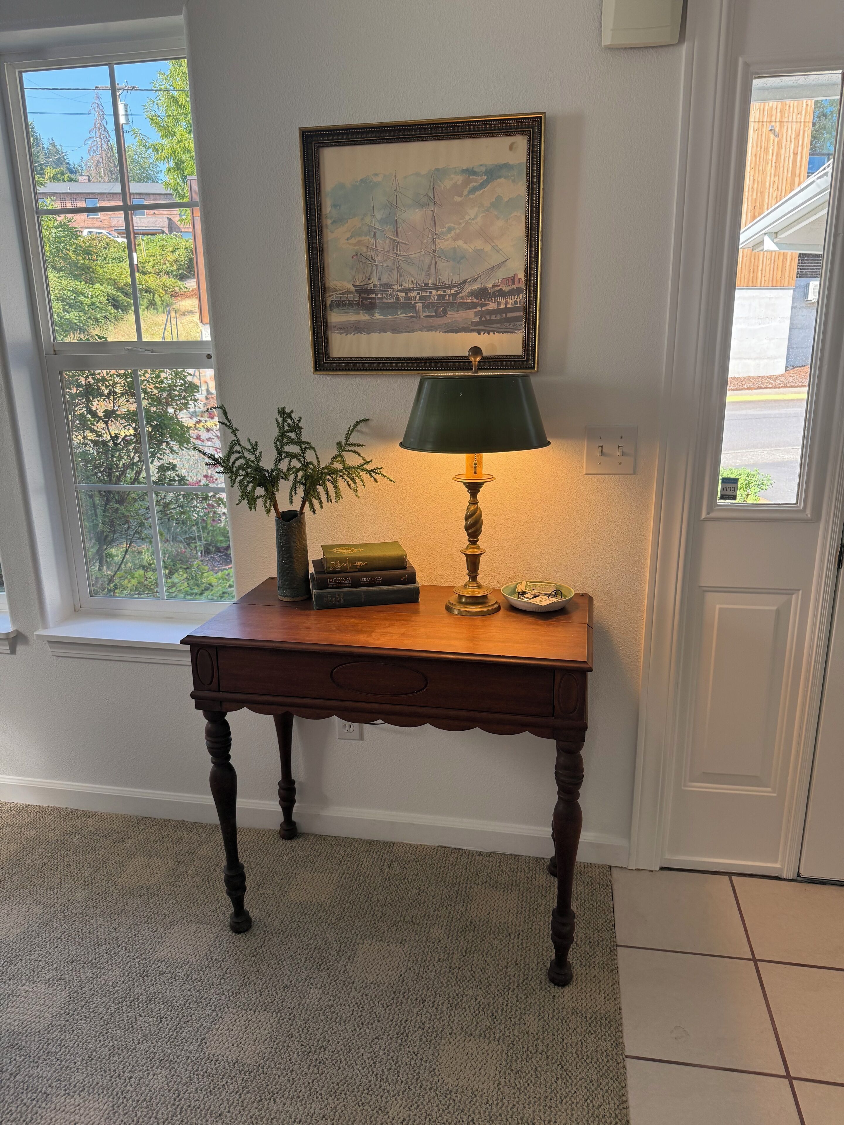 A wooden desk with a lamp, a framed painting, and a plant in a window sill.