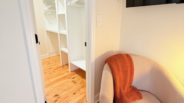 Une pièce avec un plancher en bois, une armoire blanche et un fauteuil beige avec une couverture.