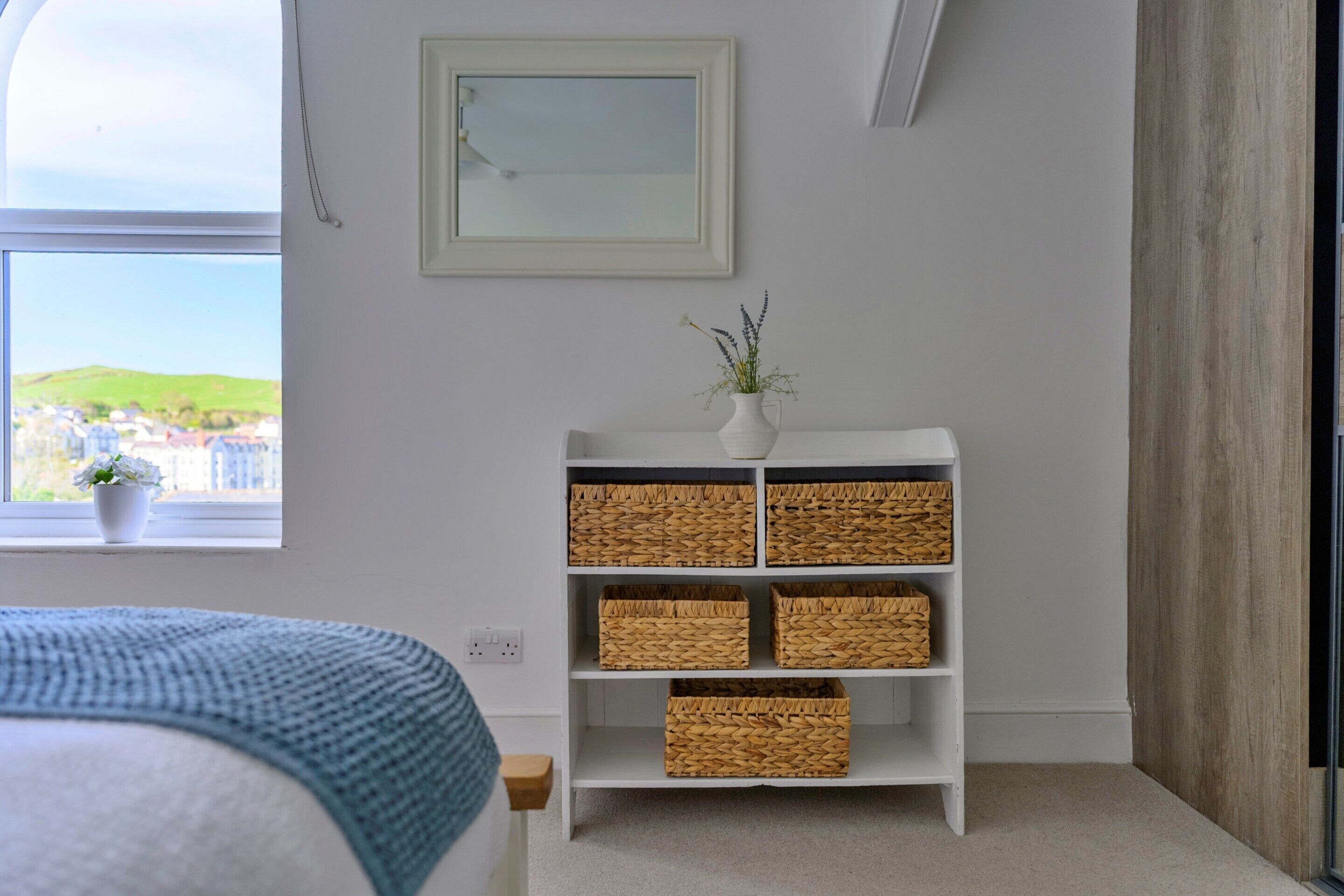 A bedroom with a bed, a white bedside table, a white wall, a window with a view, and a white shelf with baskets.