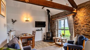 A cozy living room with a stone wall, a fireplace, a dining area, and a television. - Luxury Cornish Countryside Hut for 2 (Bude)