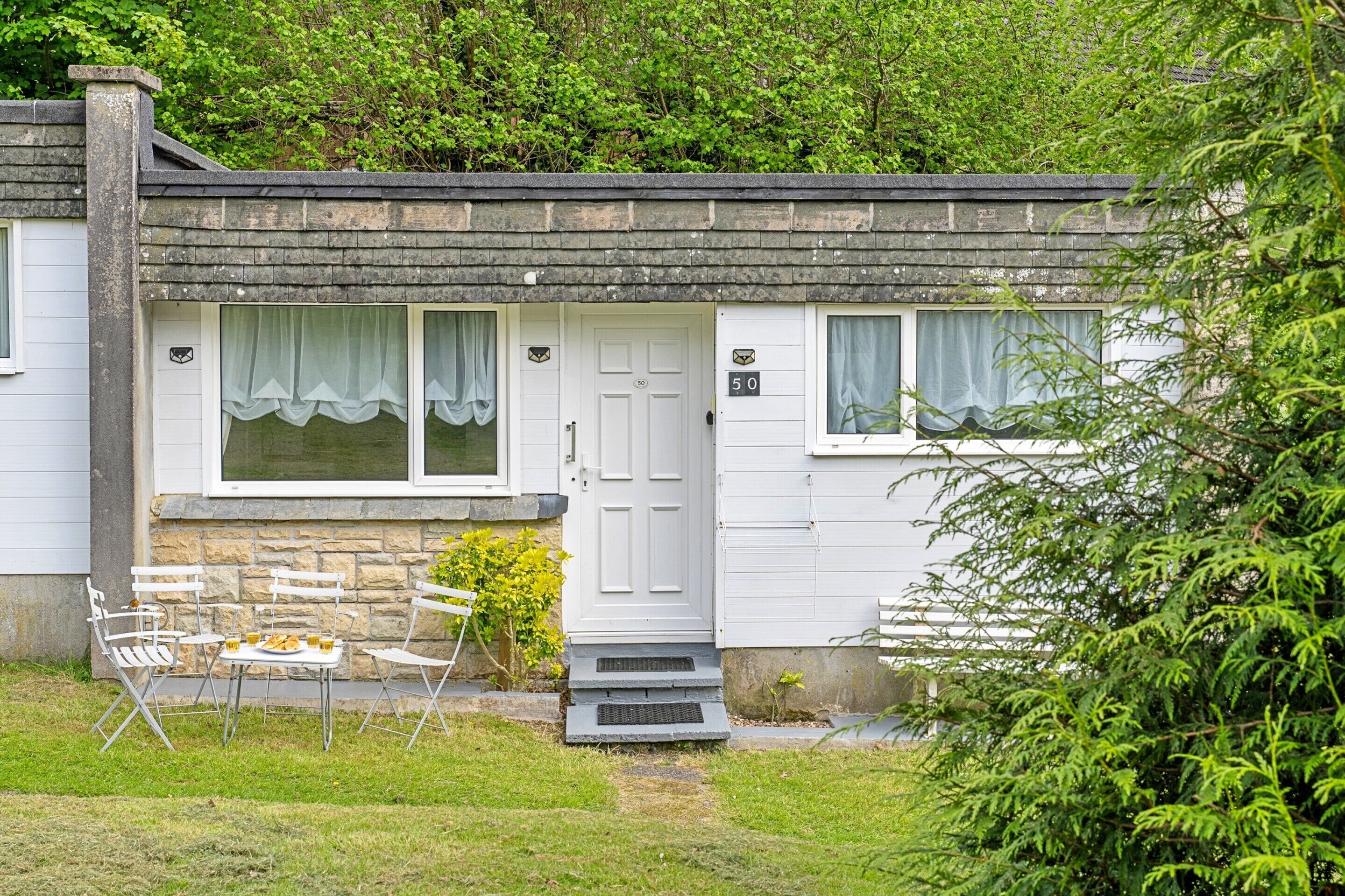 A white cottage with a stone chimney, a front door, and a small porch with a table and chairs.