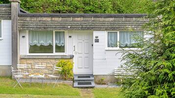 A white cottage with a stone chimney, a front door, and a small porch with a table and chairs.