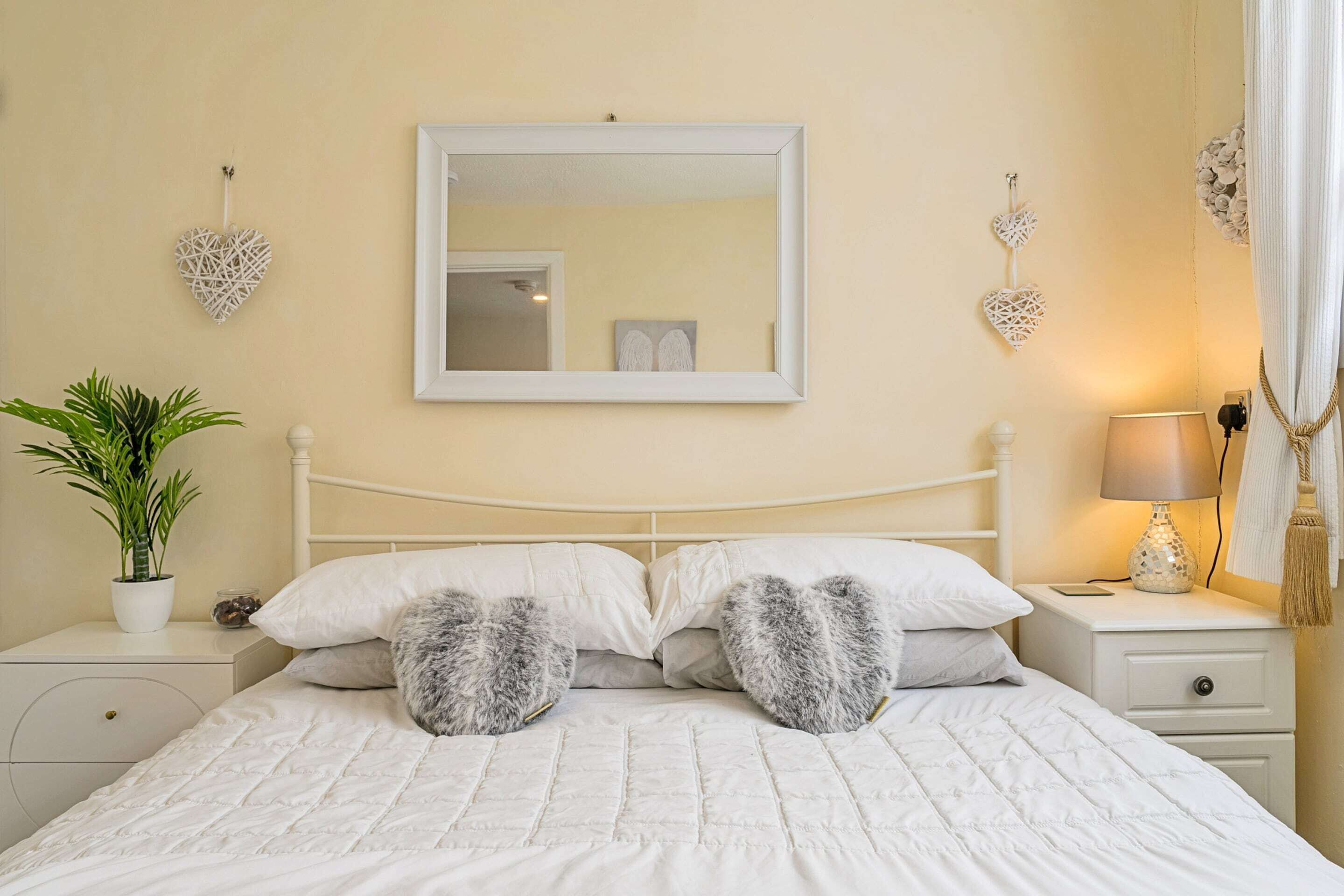 A neatly made bed with white bedding and two heart-shaped pillows, a bedside table with a lamp, a potted plant, and decorative heart-shaped wall hangings.