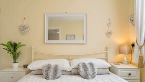 Uma cama bem arrumada com roupa de cama branca e duas almofadas em formato de coração, uma mesa de cabeceira com abajur, uma planta em vaso e elementos decorativos em forma de coração na parede.