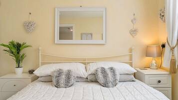A neatly made bed with white bedding and two heart-shaped pillows, a bedside table with a lamp, a potted plant, and decorative heart-shaped wall hangings.