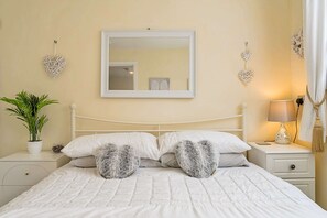 A neatly made bed with white bedding and two heart-shaped pillows, a bedside table with a lamp, a potted plant, and decorative heart-shaped wall hangings. - Camelford Base Near Beaches (Camelford)