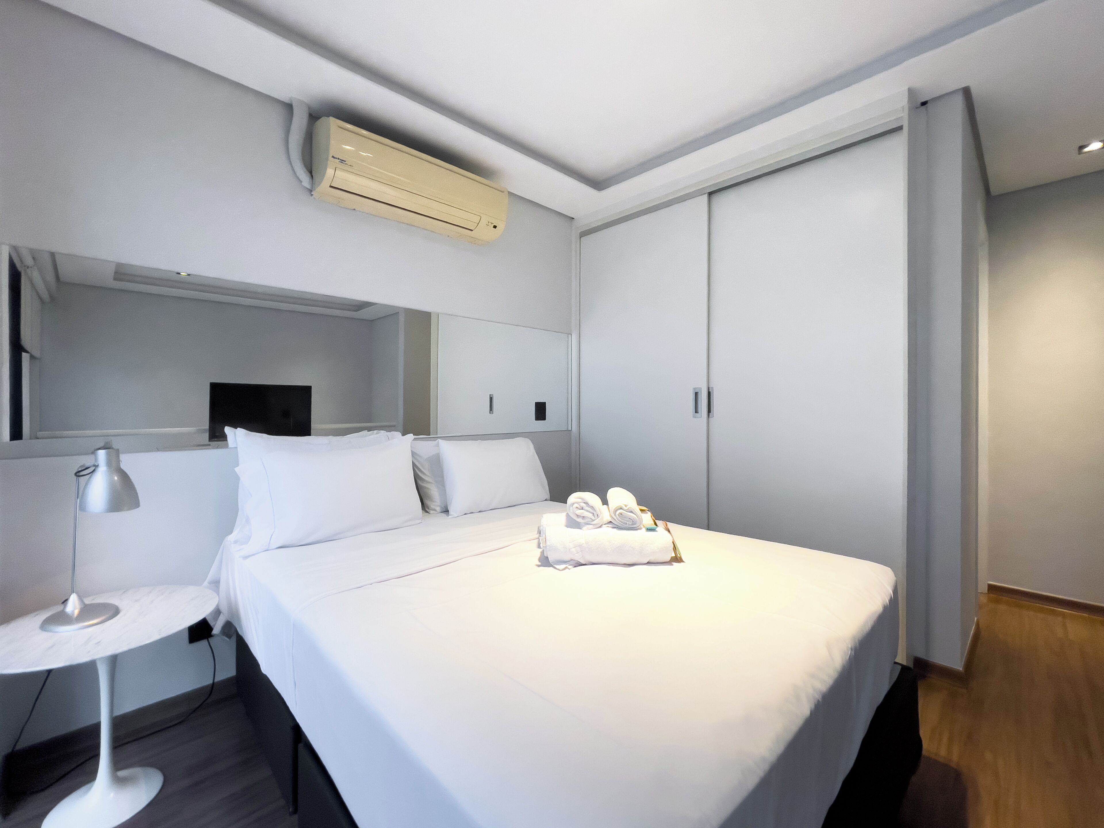 Quarto de hotel moderno com uma cama, mesa de cabeceira com abajur, espelho e guarda-roupa.