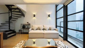 A modern living room with a sofa, a glass coffee table, a patterned rug, a staircase, and a large window with a city view.