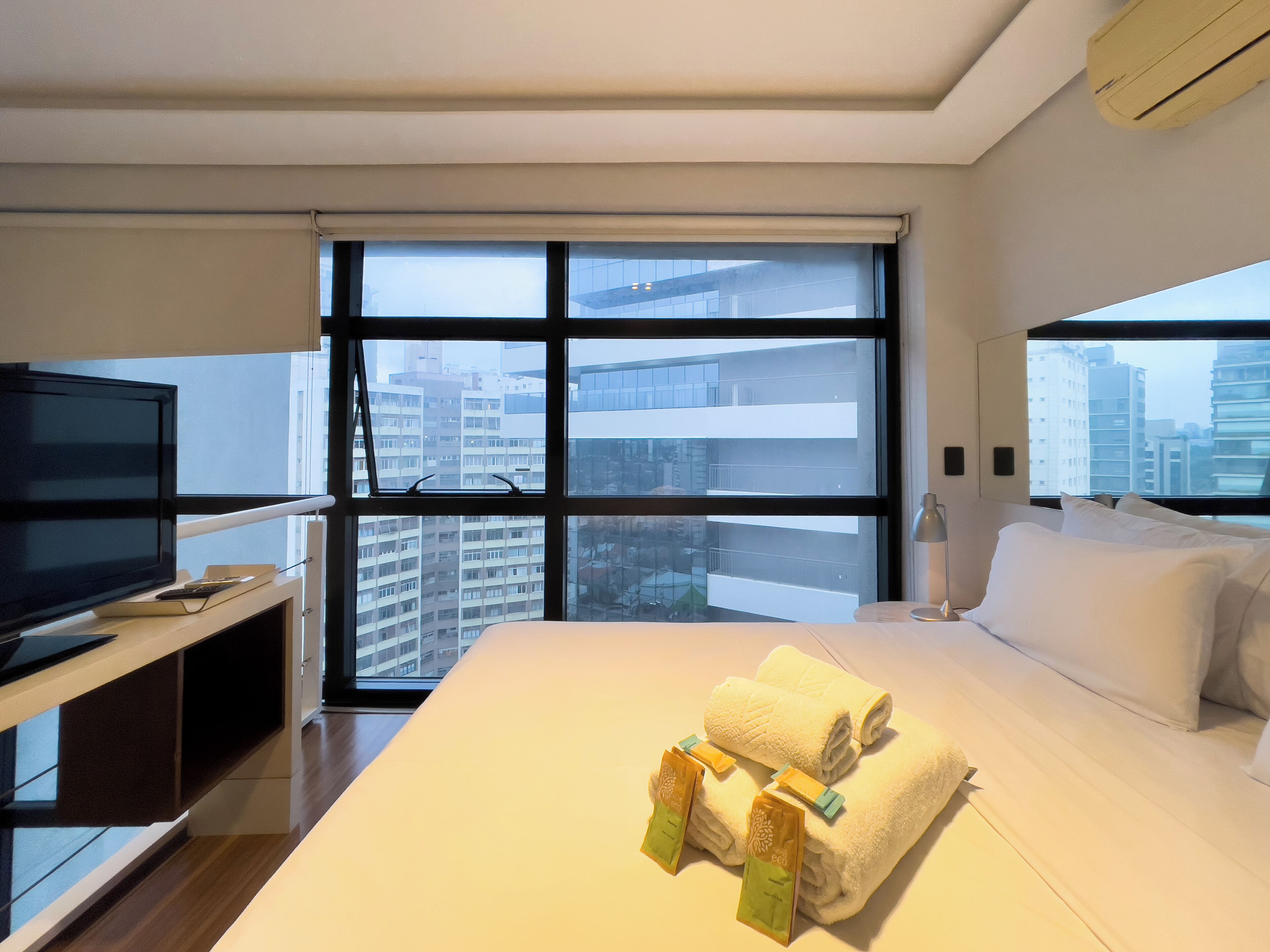 A hotel room with a large bed, a TV, and a view of the cityscape through the windows.