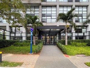 Ein Gebäudeeingang mit überdachter Veranda, Palmen und einem Sicherheitsdienstposten.