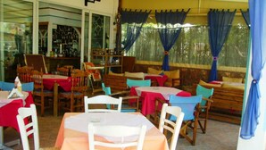 Een restaurant met meerdere tafels en stoelen, waarvan sommige rode en witte tafelkleden hebben, en een barruimte zichtbaar door een glazen scheiding.