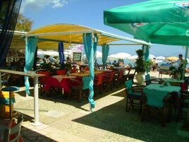 Un área de comedor al aire libre con mesas y sillas bajo una marquesina, rodeada de sombrillas.