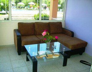 Una habitación con un sofá marrón, una mesa de centro de vidrio con libros y un jarrón de flores, y una ventana que ofrece vistas a sombrillas y sillas al aire libre.