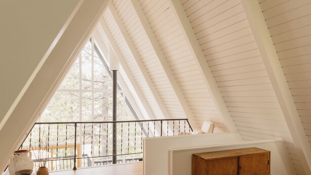 Quarto com teto inclinado, escrivaninha de madeira e uma janela ampla com vista para árvores.