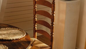 Uma mesa de jantar de madeira com um jogo americano trançado, uma cadeira de madeira com assento acolchoado e um vaso de cerâmica verde sobre um pedestal branco.