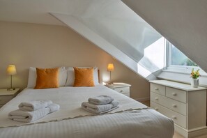 A bedroom with a sloped ceiling, a bed with white linens, two bedside lamps, and a dresser with a vase of flowers. - Charming Dorset Coach House for 6 (Fowey)