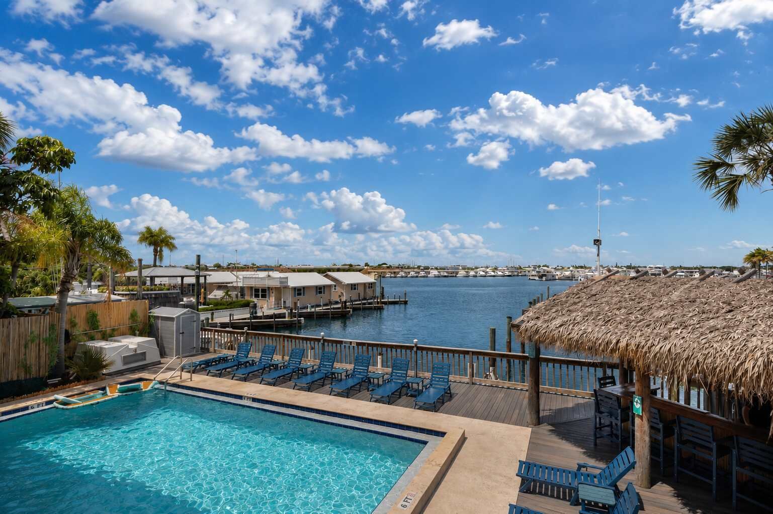A swimming pool with lounge chairs, a thatched-roof structure, and a marina in the background.