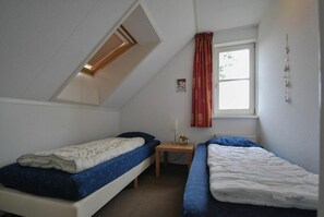 A small, single-room accommodation with two single beds, a window with curtains, a small table, and a wall-mounted light fixture. - Beachside Dune Villa With Garden (Makkum)