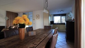 A dining area with a wooden table, chairs, and a vase of yellow flowers. There is a kitchen in the background with a window. - Beachside Dune Villa With Garden (Makkum)