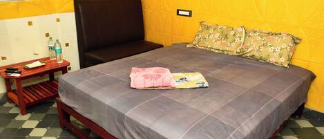 Una habitación con cama, una silla, una mesita con una botella de agua y un enchufe en la pared.