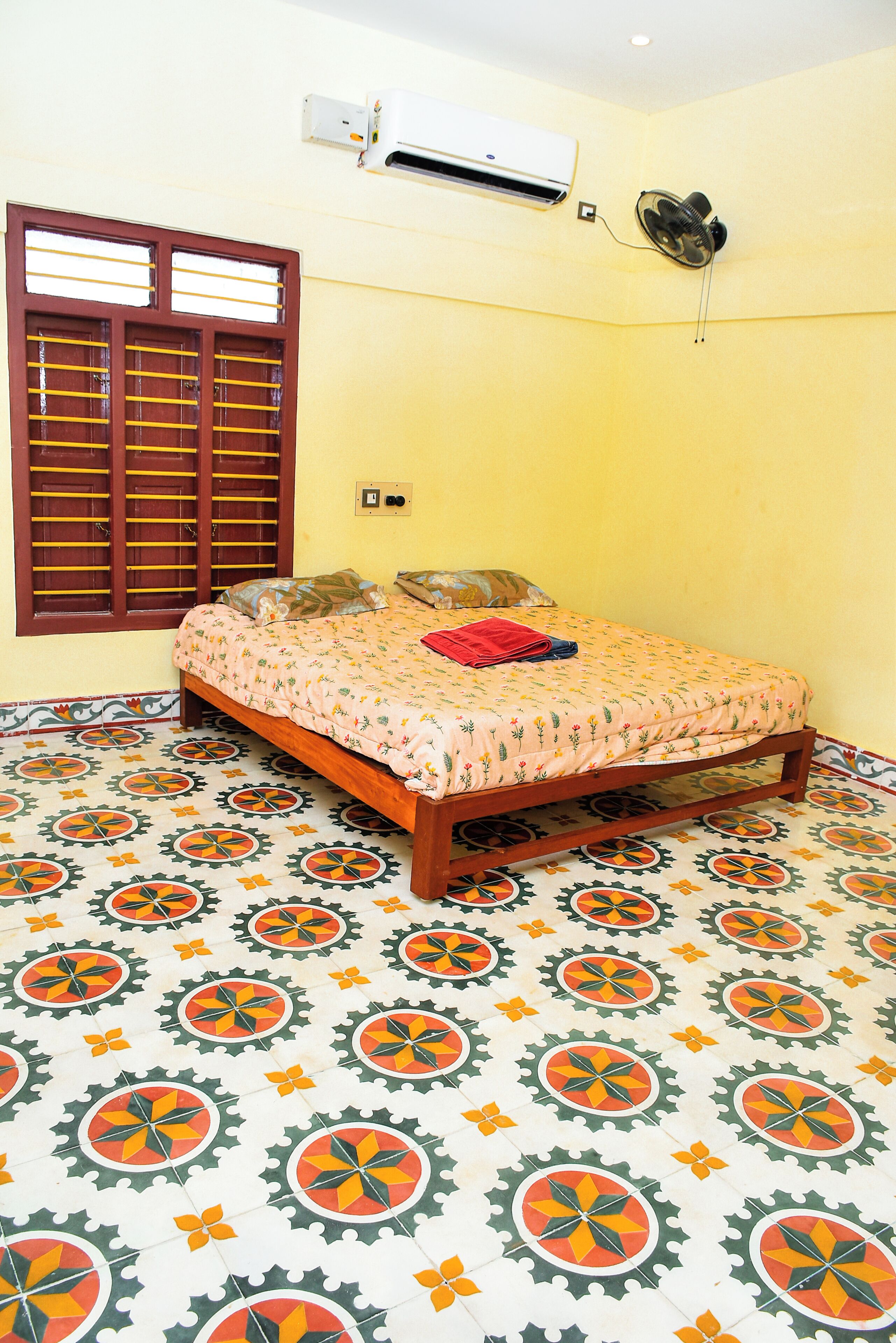 Um quarto com uma cama, ar condicionado, janela com persianas e ladrilhos no chão com padrão.