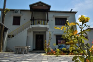 Un edificio de dos pisos con una escalera que conduce a la entrada, un banco en el exterior y una planta con flores amarillas al frente.