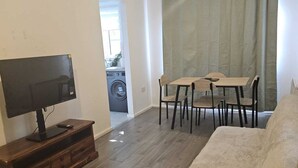 Een woonkamer met een flatscreen televisie, houten meubels, een eettafel met stoelen en een groot raam met gordijnen.