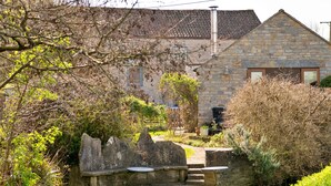 A stone building with a tiled roof, a small garden with a stone bench, and a narrow water channel. - Elegant Apartment in Regency Cheltenham (Somerton)