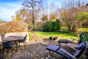 Eine Steinterrasse mit rundem Tisch und Stühlen, umgeben von einem Garten mit verschiedenen Pflanzen und einer Rasenfläche.
