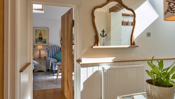 A hallway with a wooden door leading to a room with a bed and a lamp. There is a mirror on the wall and a potted plant on a table. - Modern Studio Near Port Isaac (Bodmin)