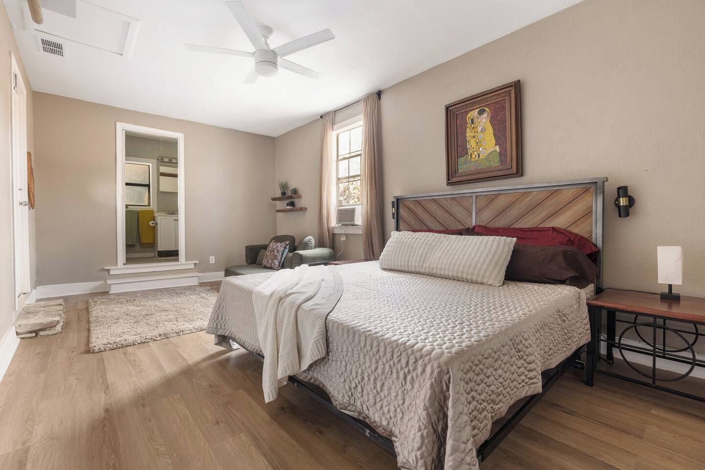 A bedroom with a bed, a nightstand, a ceiling fan, a window with curtains, a door leading to a bathroom, and a painting on the wall.