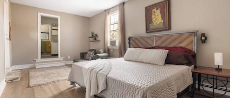 Un dormitorio con cama, mesita de noche, ventilador de techo, ventana con cortinas, puerta que da a un baño y un cuadro en la pared.