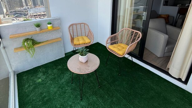 A balcony with artificial grass, two wicker chairs with cushions, a small table with a plant, and a view of the cityscape through a glass door. - Capihome (Ciudad de Guatemala)