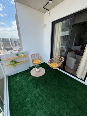 Een balkon met kunstgras, twee rieten stoelen met kussens, een kleine tafel met een plant en een uitzicht op de stad door een glazen deur.