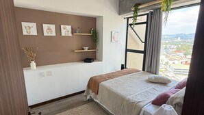 A bedroom with two beds, a window with a city view, a wall-mounted shelf with decorative items, and a ceiling light fixture. - Capihome (Ciudad de Guatemala)