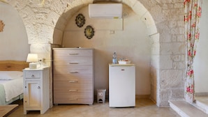 Ein Zimmer mit einem Steingewölbe, einem Holzkleiderschrank, einem kleinen weißen Kühlschrank und einem Bett mit einem hölzernen Kopfteil.