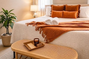 A neatly made bed with a brown blanket and pillows, a small wooden table with a candle, and a potted plant. - 2-Home Austin Compound | Hot Tub + Cowboy Pool (Austin)