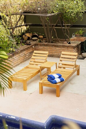 A poolside area with wooden lounge chairs and a small table, surrounded by greenery and a brick wall. - Beautiful home in La Punta with a private pool, rooftop terrace, and AC. (Puerto Escondido)