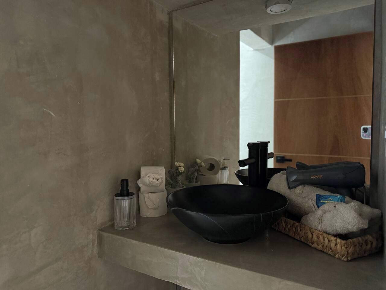 A bathroom with a large mirror, a black basin, a towel dispenser, and a small statue.