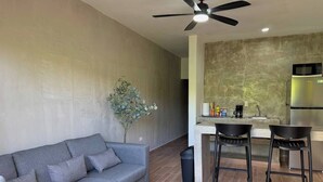 Um espaço de estar moderno com um sofá cinzento, uma pequena cozinha com micro-ondas e um ventilador de teto.