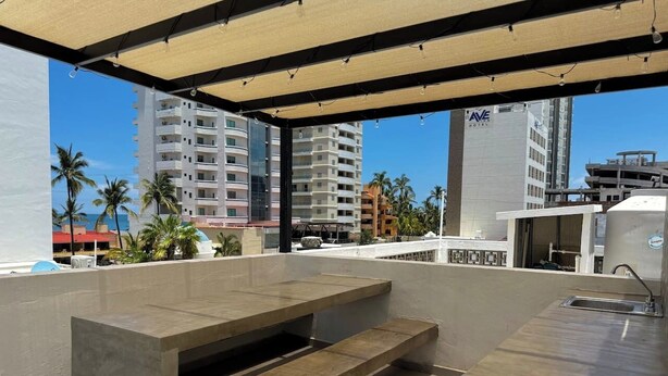 Uma área de pátio ao ar livre com mesa e bancos de concreto, uma pia de metal e vista para palmeiras e edifícios.