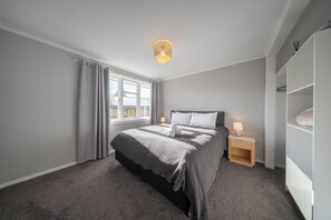 Ein Schlafzimmer mit Bett, Nachttisch, Lampe, Kleiderschrank und einem Fenster mit Vorhängen.