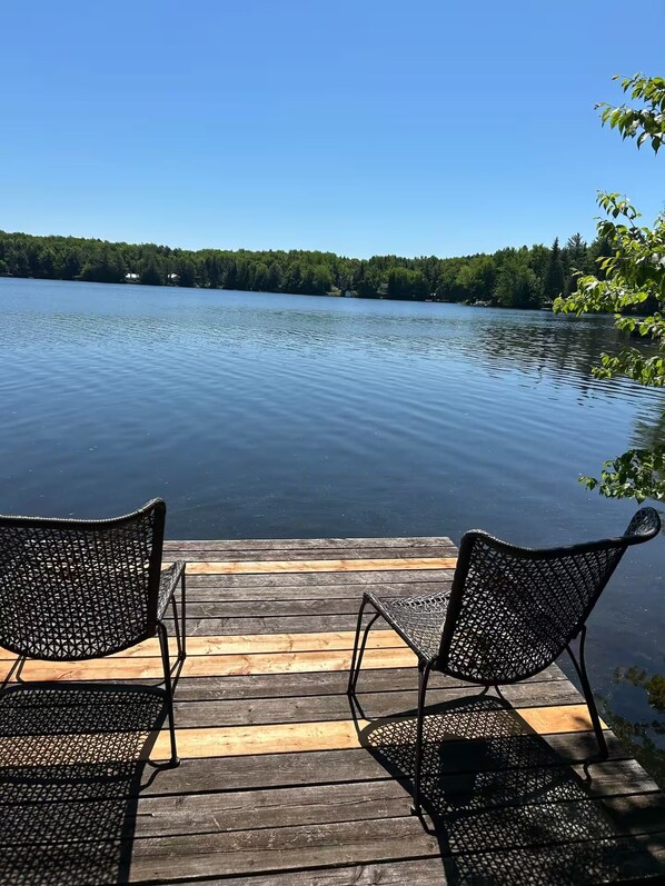 A wooden dock with two metal chairs facing a calm lake, surrounded by trees.