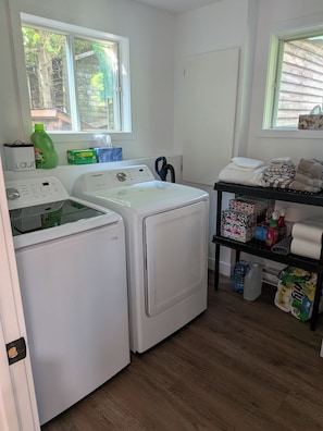 A laundry room with two white front-loading washers and dryers, a shelf with cleaning supplies, and a window with a view of trees. - Lakeside Chalet Welcomes You — Experience tranquility in nature. Pet-friendly! (Saint-Étienne-de-Bolton)
